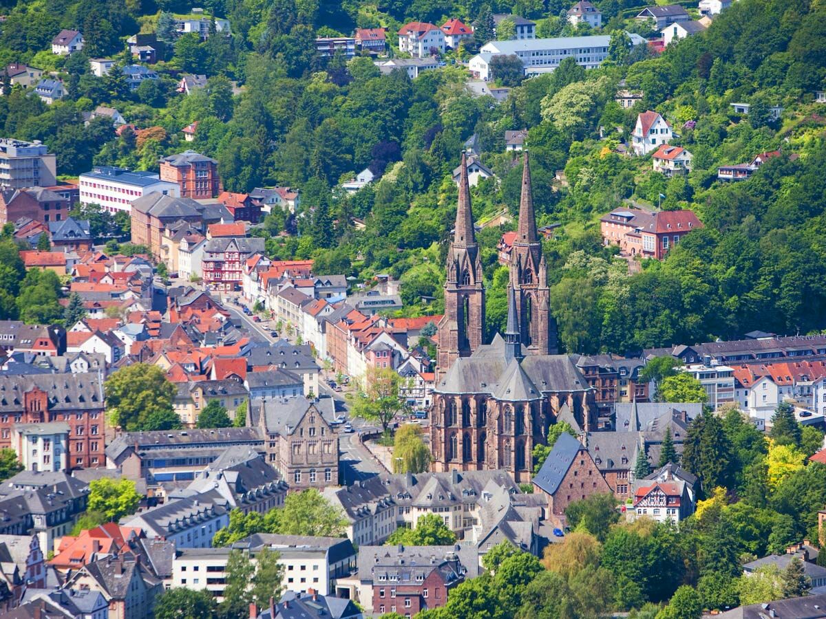 Luftaufnahme von Marburg mit der Elisabethkirche und vielen Häusern auf grünen Hügeln unter blauem Himmel.
