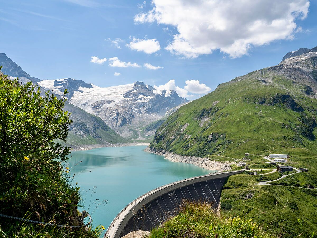 Türkisfarbener Gletschersee mit großem Staudamm, umgeben von grünen Berghängen und schneebedeckten Alpen in Österreich.