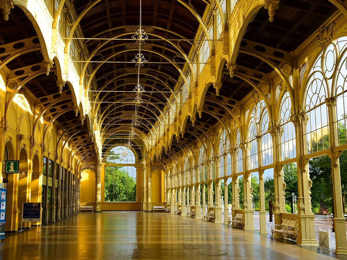 Elegante, lange goldene Kolonnaden-Interieur mit Bogenfenstern, verzierten Details und glänzendem Boden, mit Blick auf einen grünen Park.