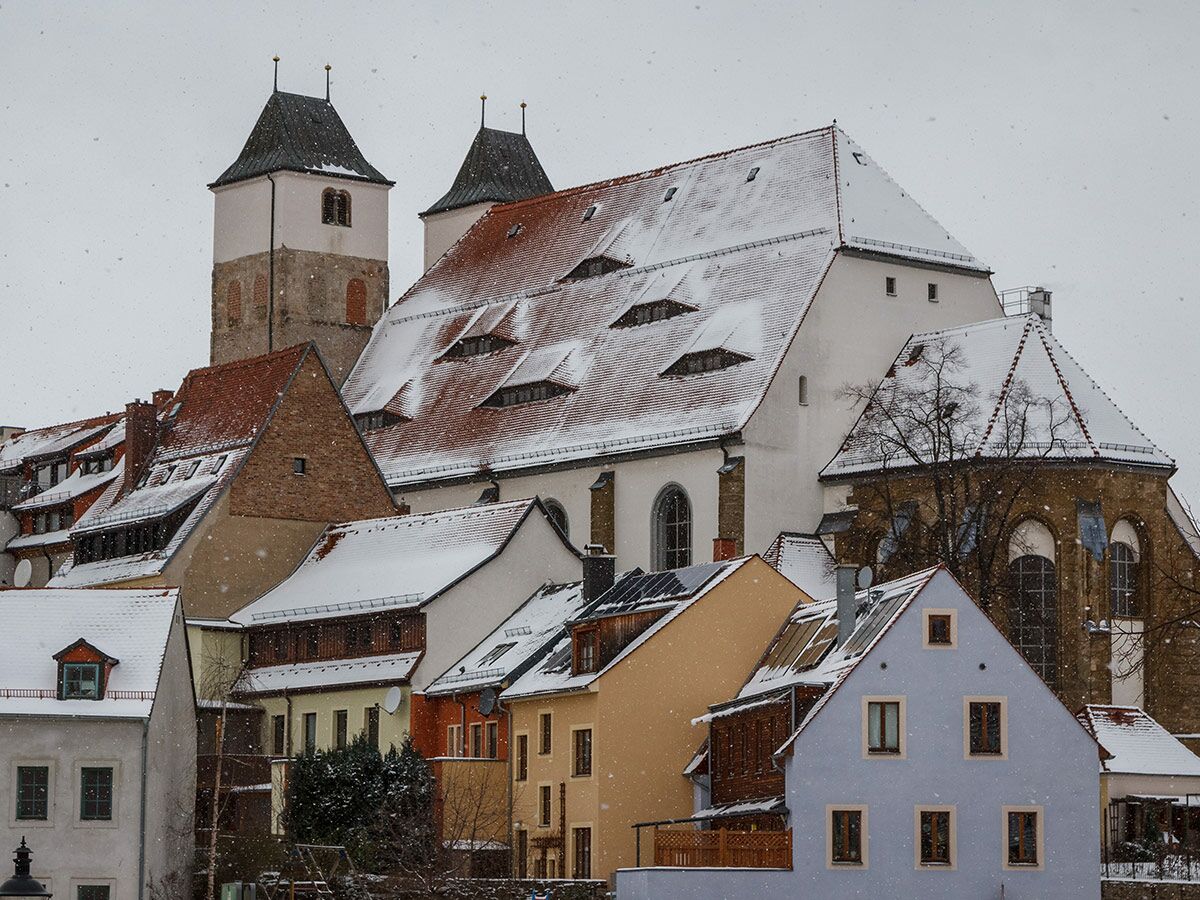Winterliche Stadtansicht mit schneebedeckten Dächern, Kirchtürmen und bunten Häusern unter leichtem Schneefall.