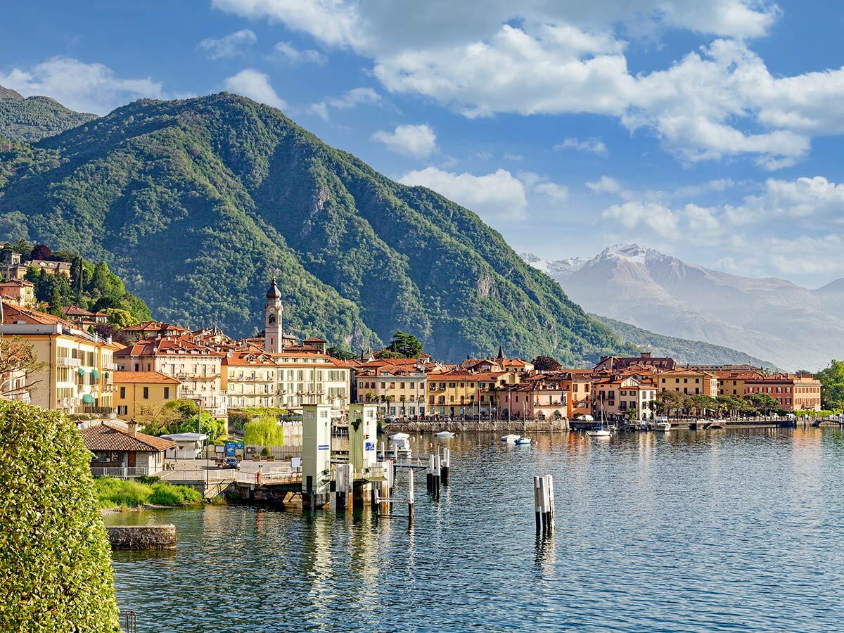 Malerische italienische Stadt mit bunten Häusern und Kirchturm am Ufer eines blauen Sees, umgeben von grünen Bergen und schneebedeckten Gipfeln.