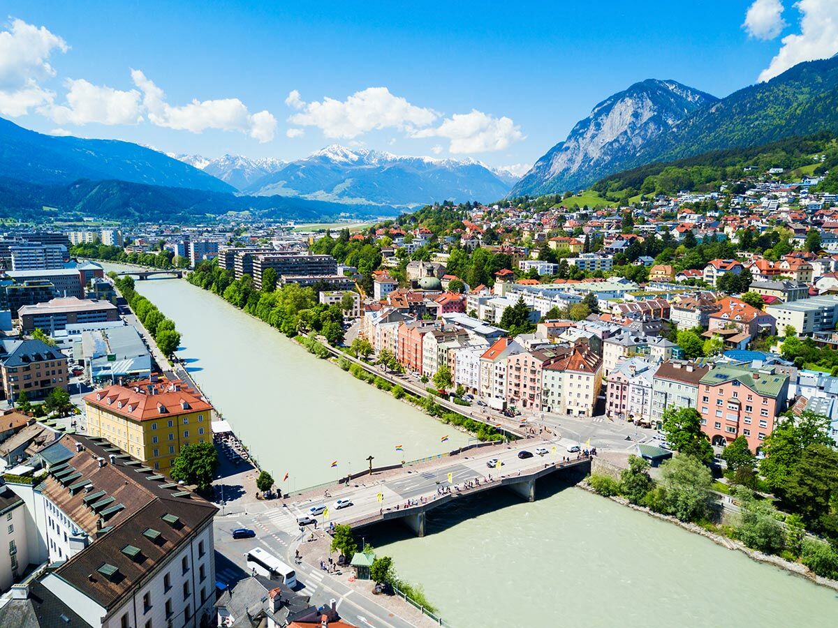 Luftaufnahme von Innsbruck mit dem Fluss Inn, farbenfrohen Gebäuden, Brücken und den umliegenden Alpen unter blauem Himmel.