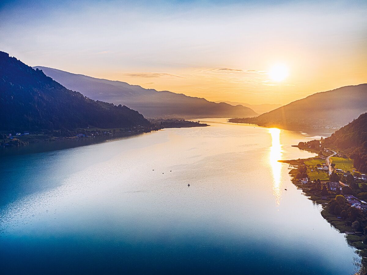 Panoramablick auf einen Bergsee bei Sonnenuntergang mit spiegelndem Sonnenlicht und umliegenden Bergen.