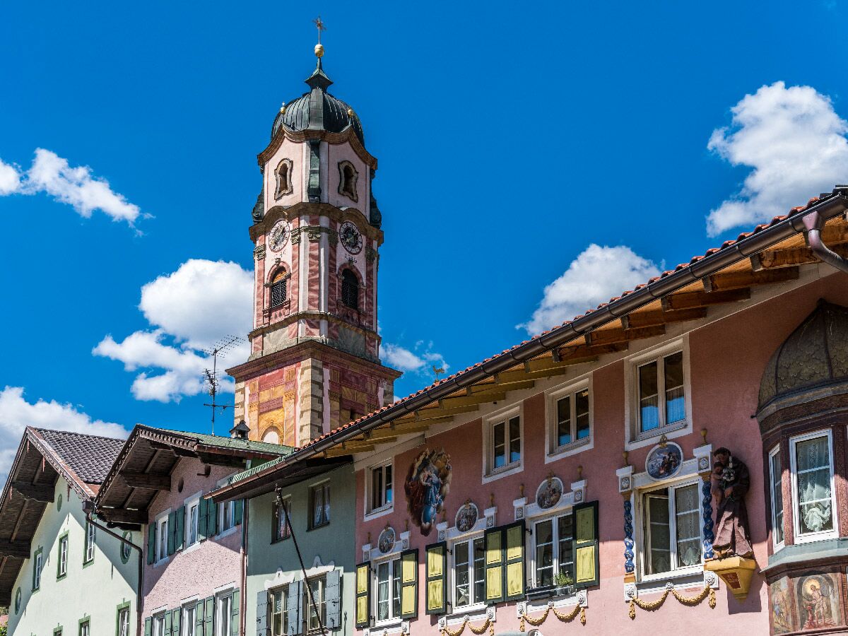 Detailreicher Barockkirchturm und traditionelle, farbenfrohe Häuser in einer bayerischen Alpenstadt am sonnigen Tag.