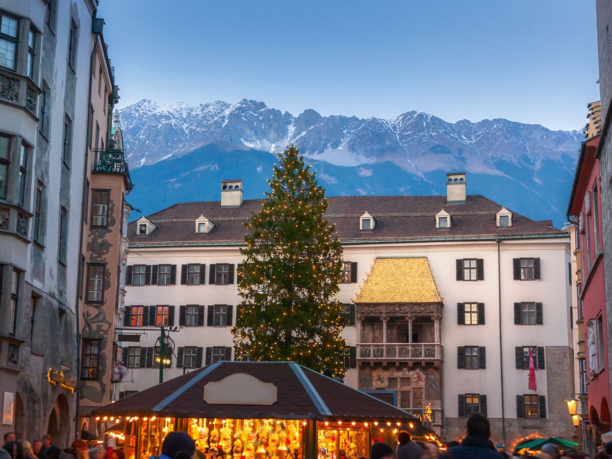 Abendaufnahme des Weihnachtsmarkts in Innsbruck mit beleuchtetem Goldenen Dachl, Christbaum und schneebedeckten Alpen im Hintergrund.