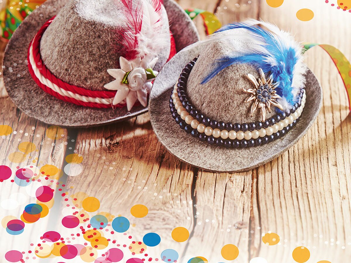 Zwei traditionelle bayerische Trachtenhüte mit Federn und Edelweiß auf Holztisch, umgeben von buntem Konfetti, symbolisieren Festlichkeit.