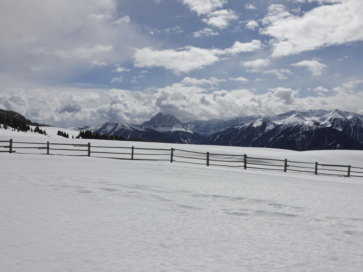 Panoramablick auf eine winterliche Alpenlandschaft mit schneebedeckten Bergen, einem hölzernen Zaun im Vordergrund und dramatischen Wolken.