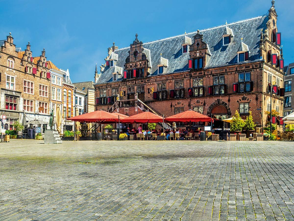 Ein sonniger historischer Marktplatz mit alten Backsteingebäuden, Restaurants mit roten Schirmen und Menschen.