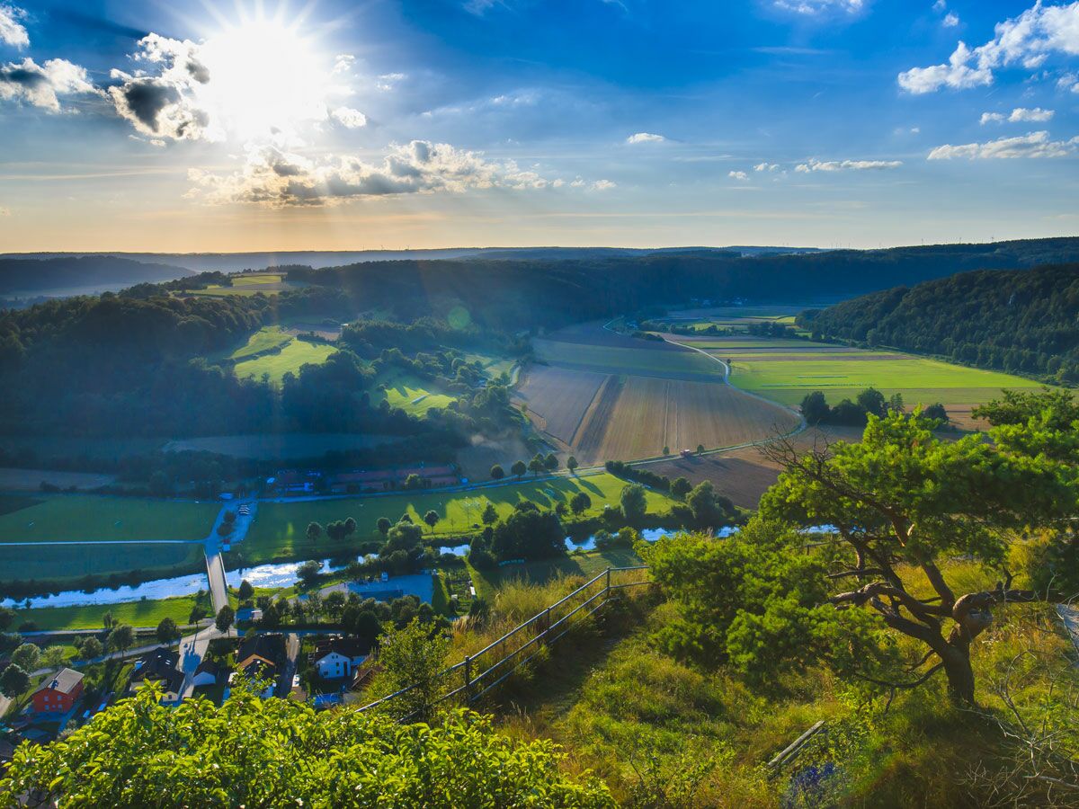 Panoramablick auf ein Flusstal bei Sonnenuntergang mit grünen Feldern, Wäldern, einem Dorf und einer Brücke unter strahlendem Himmel.