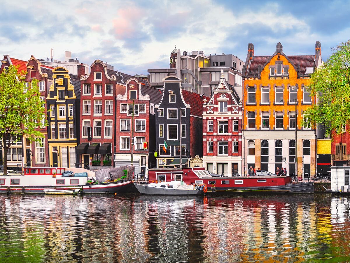Bunte Grachtenhäuser in Amsterdam, darunter Boote und ihre Reflexionen im Kanal unter einem bewölkten Himmel.