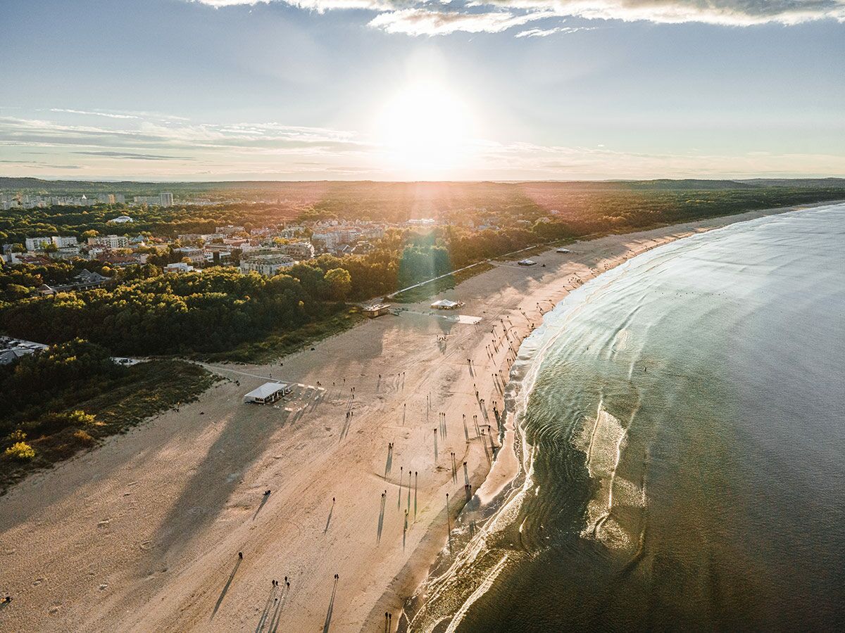Luftaufnahme vom Strand bei Sonnenuntergang mit langen Schatten von Menschen, dem Meer und einer dahinterliegenden Stadt.