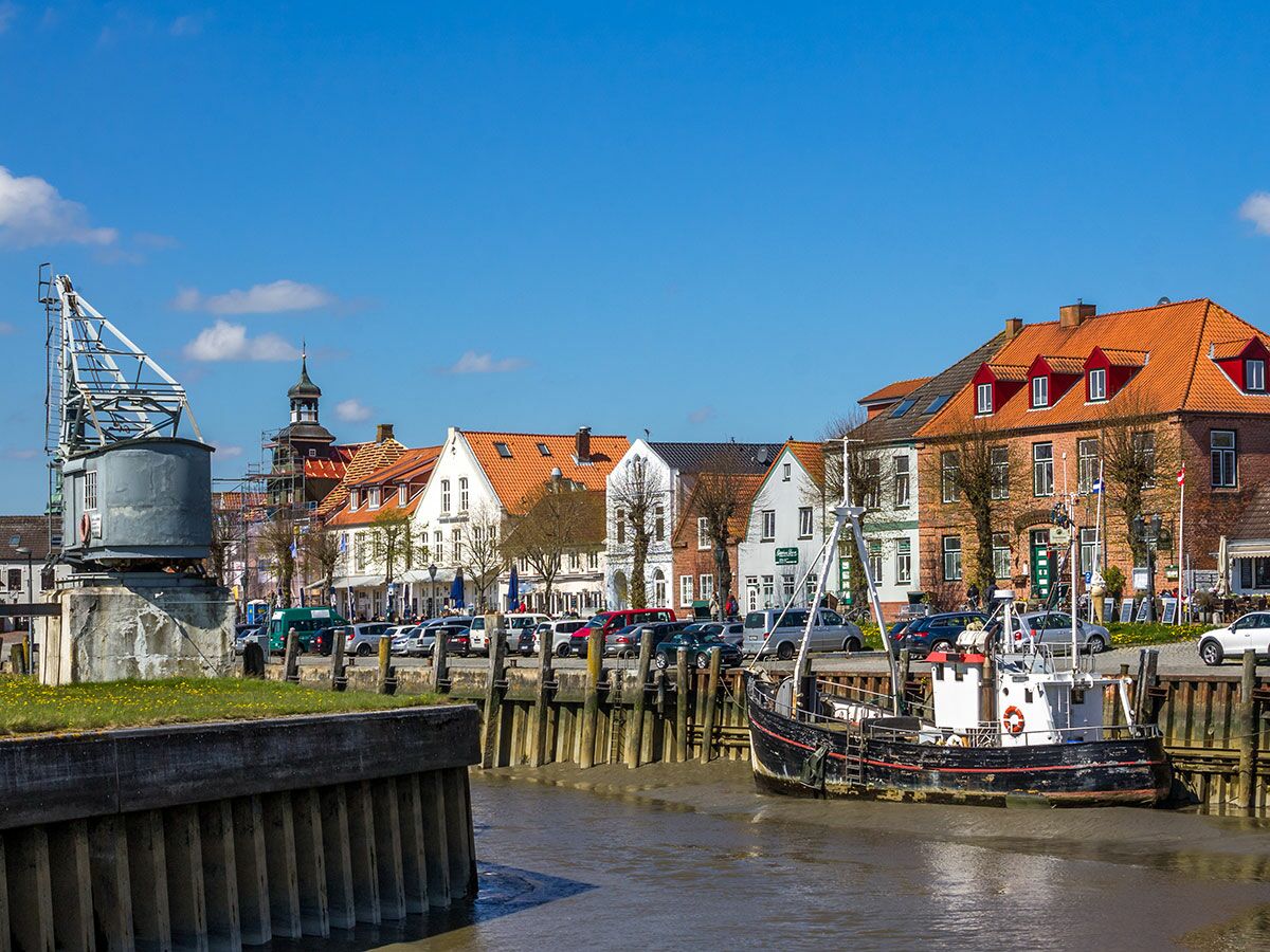 Blick auf eine historische Hafenstadt mit bunten Häusern, einem alten Fischerboot und einem Kran am Kanalufer bei sonnigem Wetter.