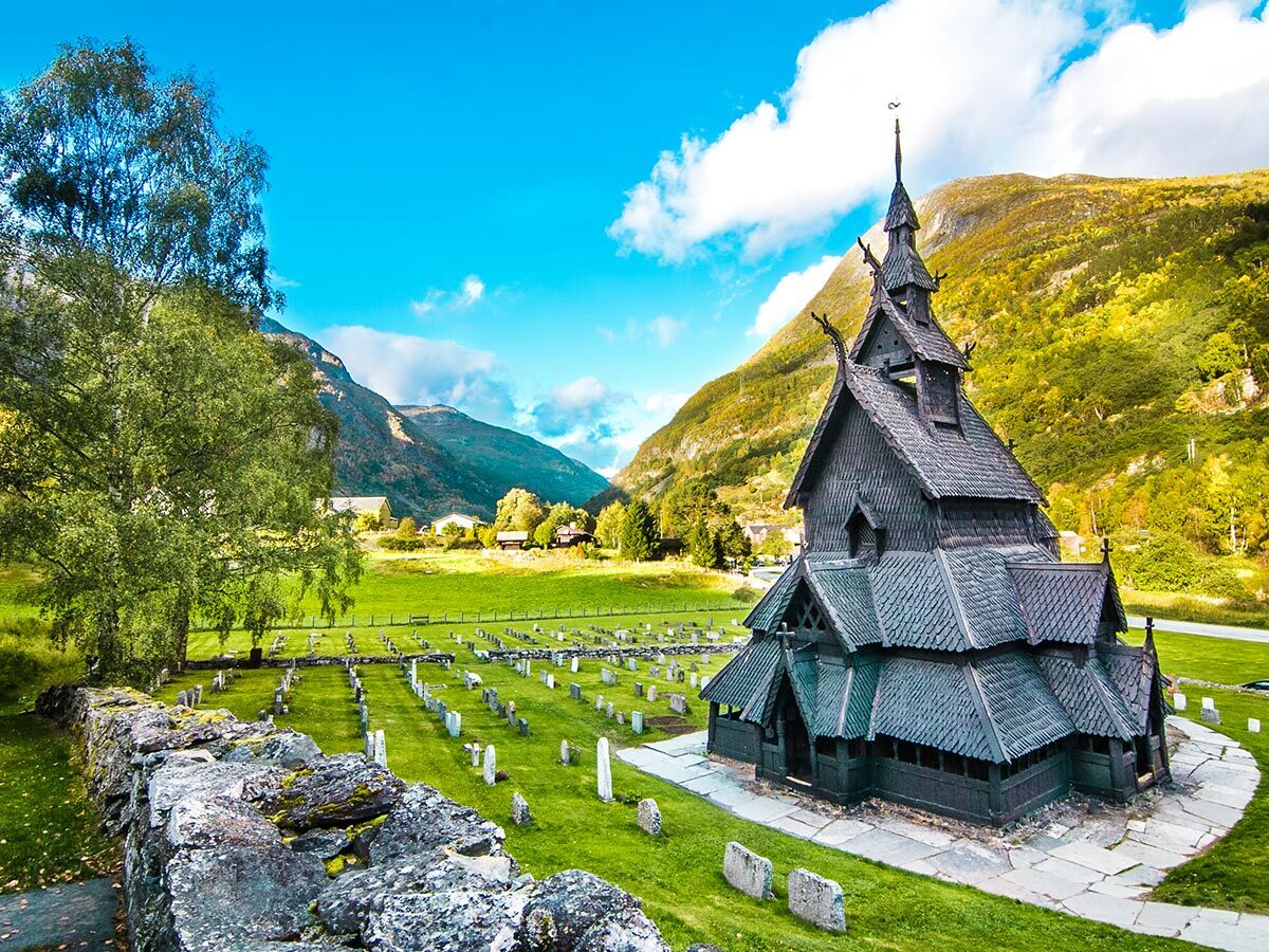 Historische Borgund Stabkirche in Norwegen mit Friedhof und Bergpanorama unter blauem Himmel.
