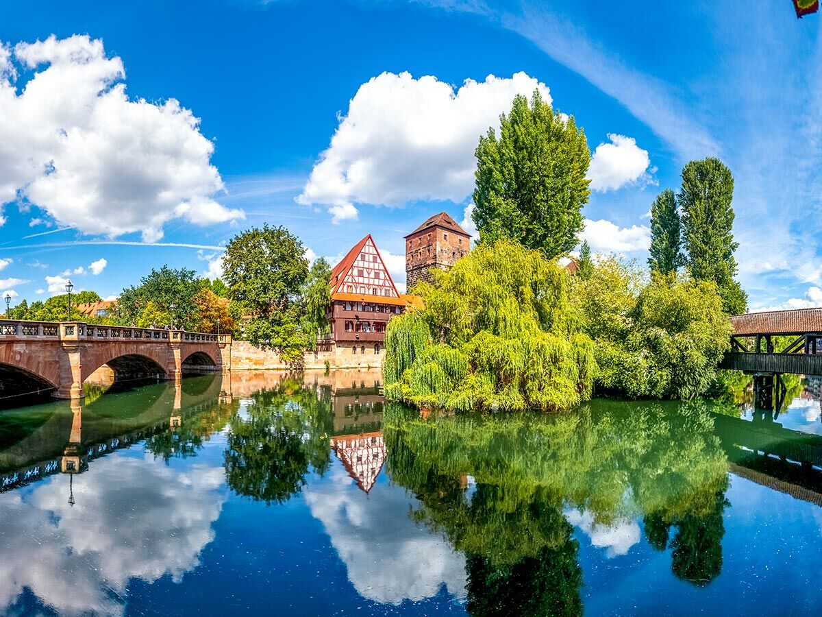 Nürnberger Altstadtansicht mit Pegnitz, historischer Steinbrücke, Fachwerkhäusern, Burgturm und Holzkontorbrücke bei Sonnenschein.