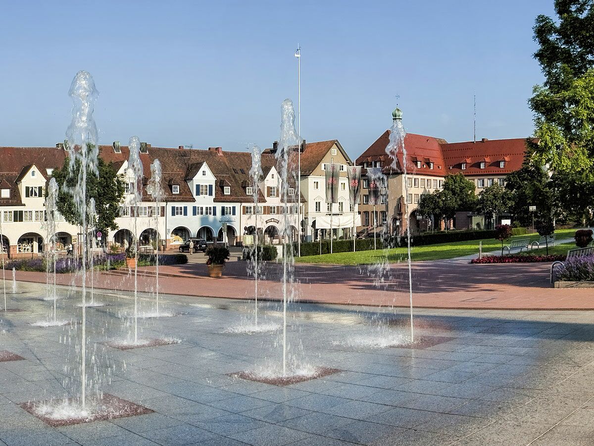 Moderner Stadtplatz mit Bodenbrunnen, charmanten europäischen Gebäuden, roten Dächern und Grünflächen unter blauem Himmel.