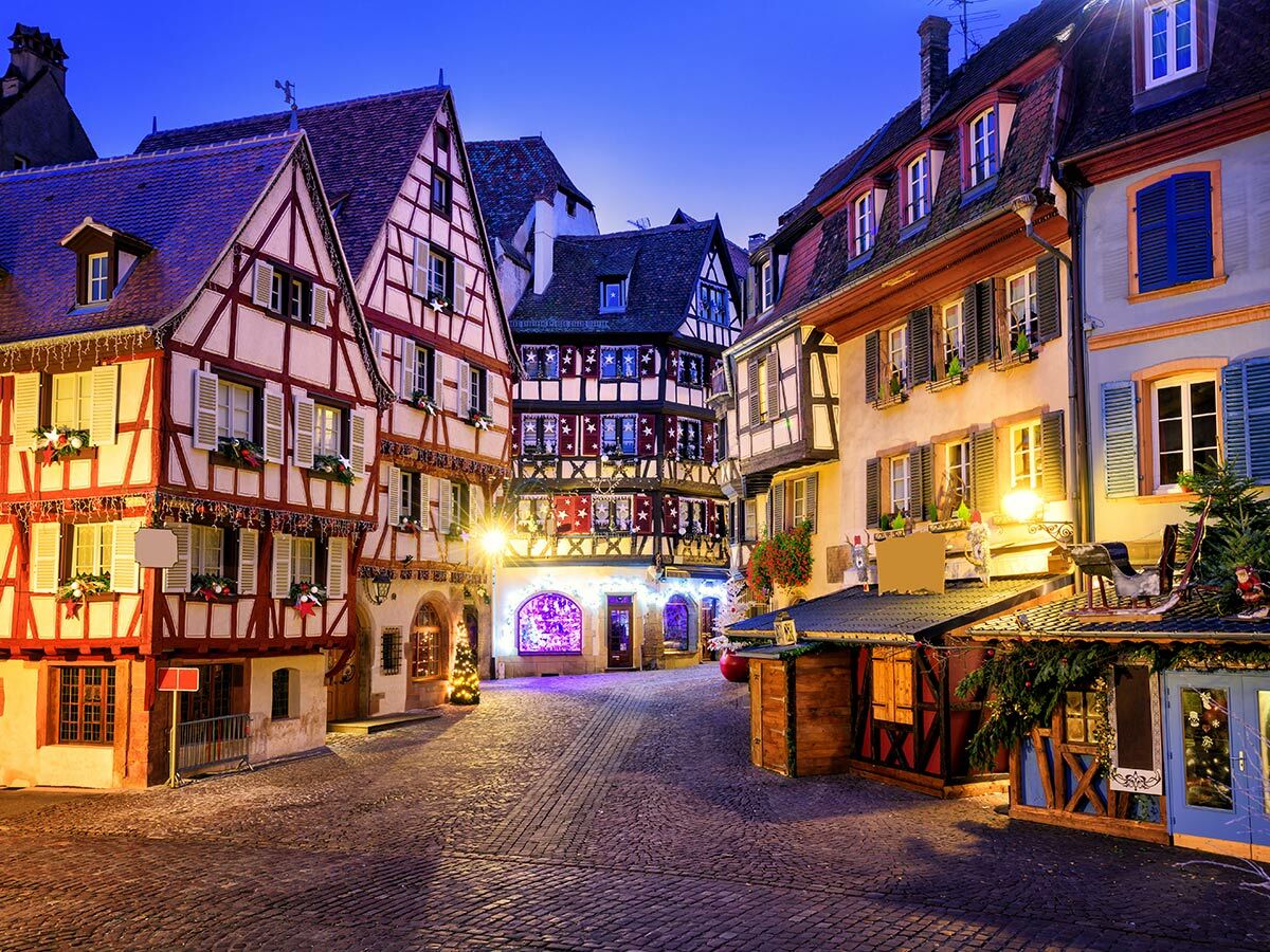 Festlich beleuchtete Fachwerkhäuser in Colmar bei Nacht mit Weihnachtsdekoration und Marktständen auf Kopfsteinpflaster.