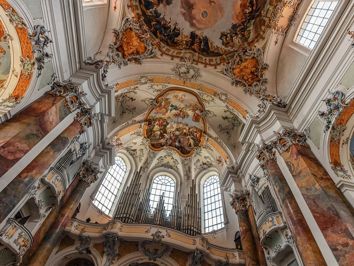 Blick in eine prächtige Barockkirche mit verzierten Säulen, Deckenfresken, goldenem Stuck und einer großen Orgel.