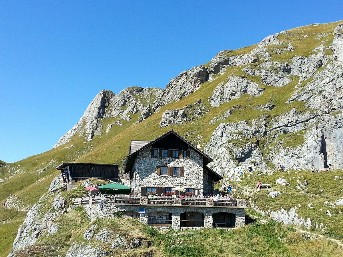 Traditionelle Berghütte aus Stein mit Terrasse auf einem grünen Berghang unter blauem Himmel. Wanderer genießen die Sonne.