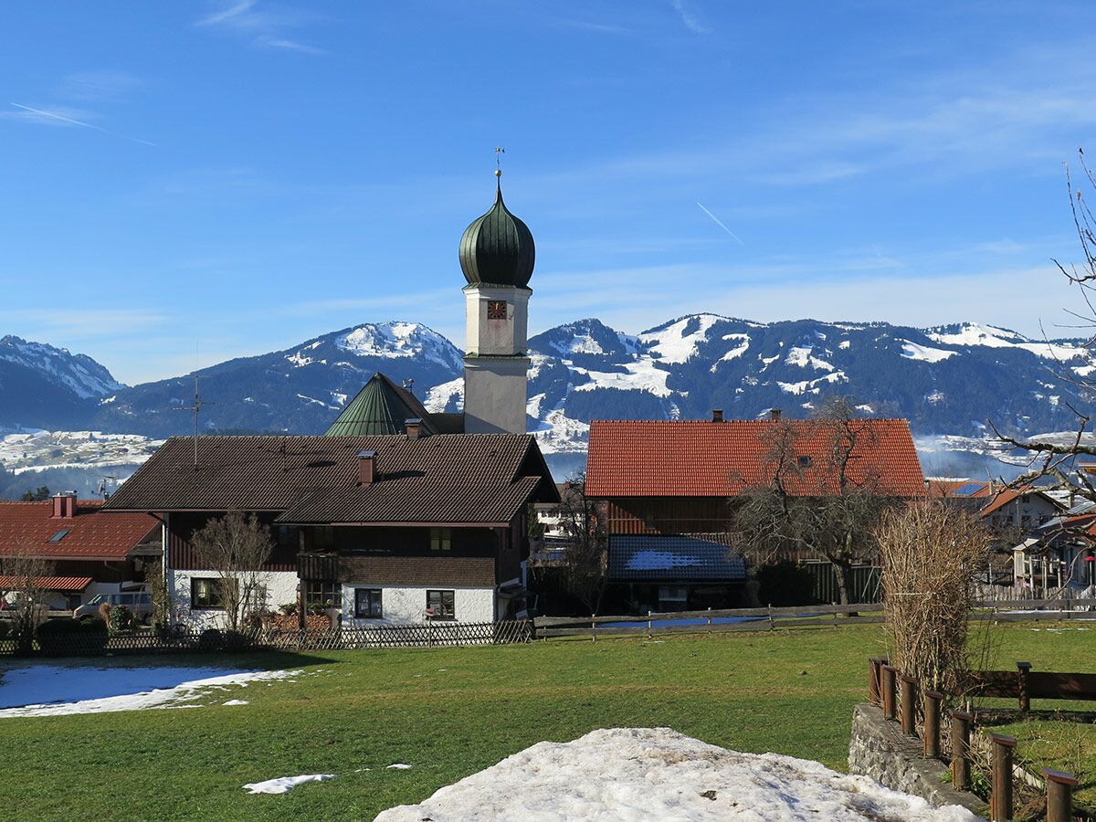 Malerisches Alpendorf mit traditionellen Häusern und einer Kirche mit Zwiebelturm vor schneebedeckten Bergen unter blauem Himmel.