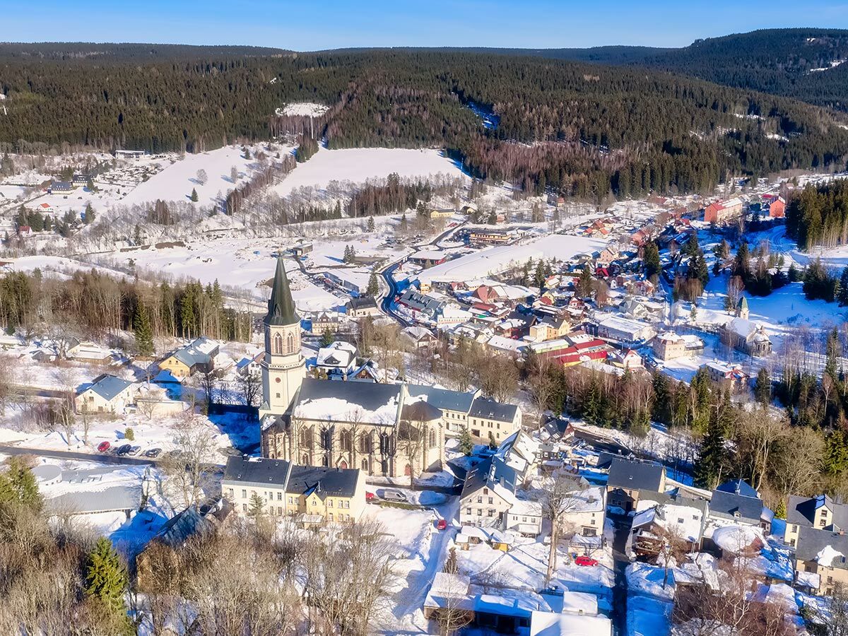 Luftaufnahme eines verschneiten Bergdorfes mit markanter Kirche, umgeben von dichten Winterwäldern und schneebedeckten Häusern.