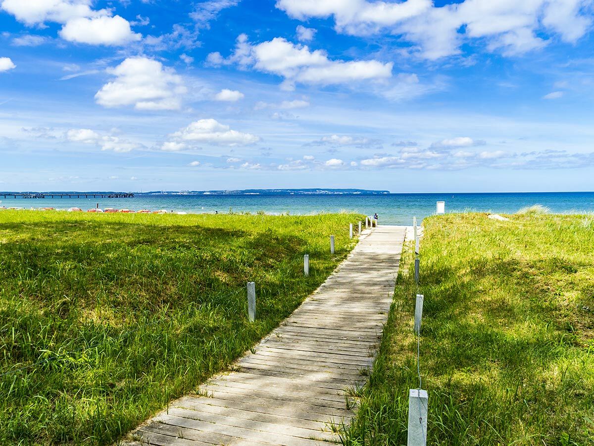 Ein sonniger Tag am Strand mit einem Holzsteg, der durch grüne Dünen zum blauen Meer und einem langen Pier führt.