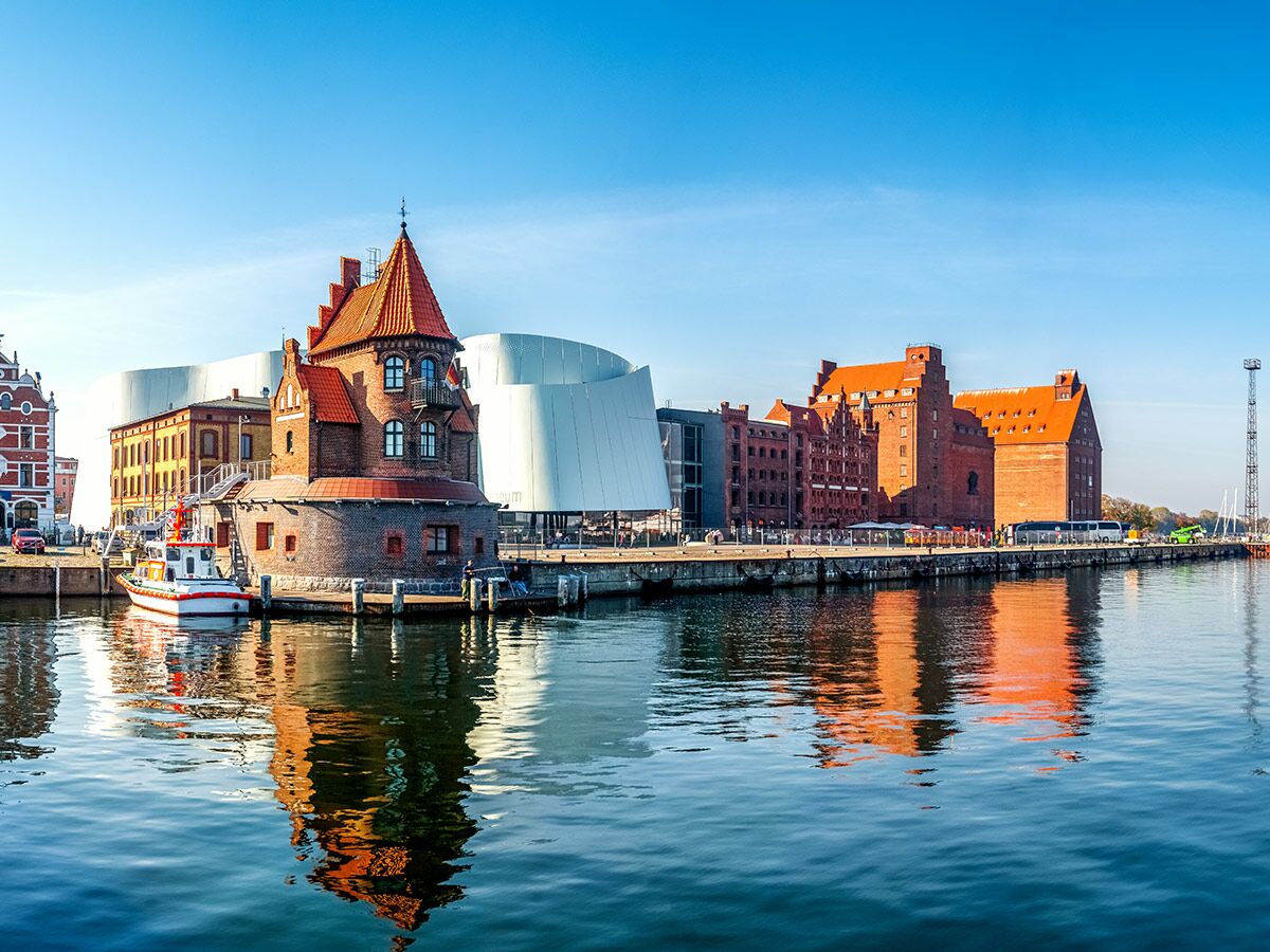 Panoramablick auf den Hafen von Stralsund mit historischem Zollhaus, dem Ozeaneum und Backsteingebäuden, die sich im Wasser spiegeln.