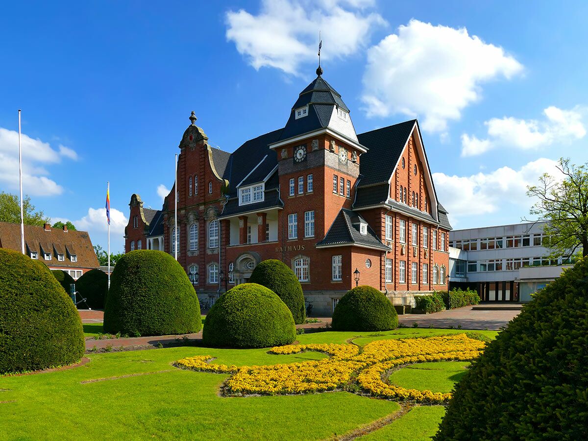 Großes rotes Backstein-Rathaus mit Turm, Uhr und geschwungenem gelbem Blumenbeet auf grünem Rasen unter blauem Himmel.