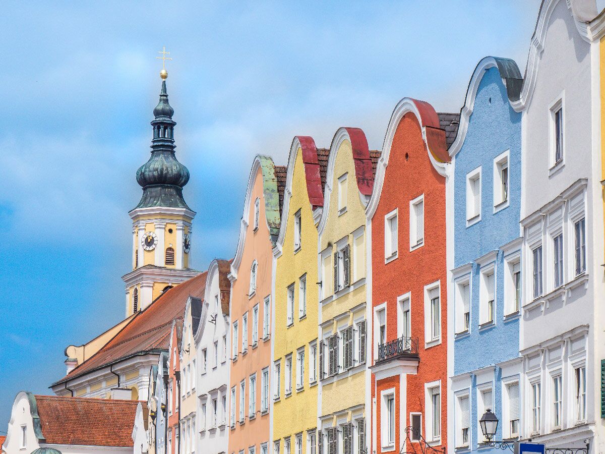 Reihe bunter Häuser mit geschwungenen Giebeln und barocker Kirchturm mit Zwiebelhaube und Uhr vor blauem Himmel.