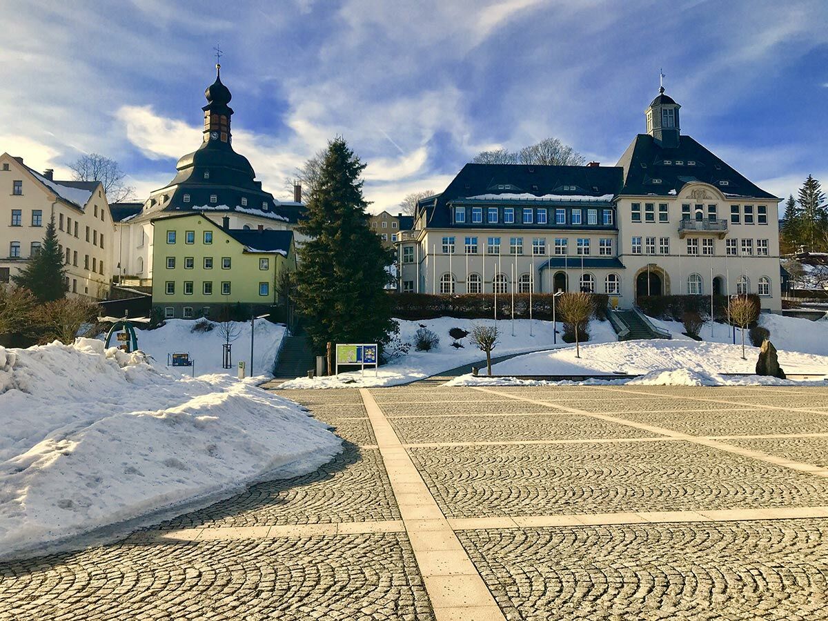Ein winterlicher Stadtplatz mit Kopfsteinpflaster, Schnee, einer Kirche mit Zwiebelturm und einem historischen Rathaus unter blauem Himmel.