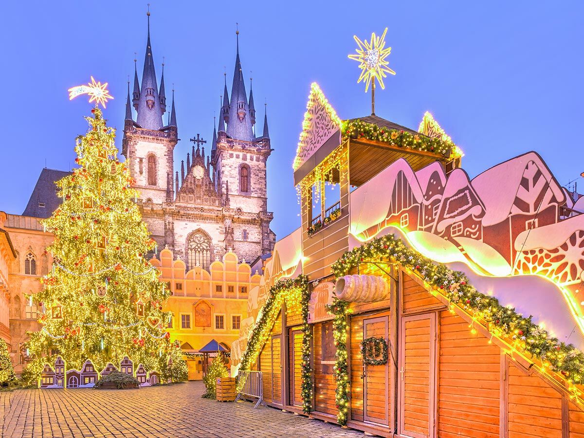 Festlicher Prager Weihnachtsmarkt bei Dämmerung mit hell erleuchtetem Christbaum, Tyn-Kirche im Hintergrund und geschmückten Buden.