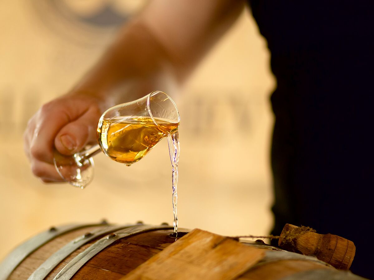 Eine Hand gießt goldfarbenen Whisky aus einem Glas in ein Holzfass; Qualitätsprüfung in Destillerie.