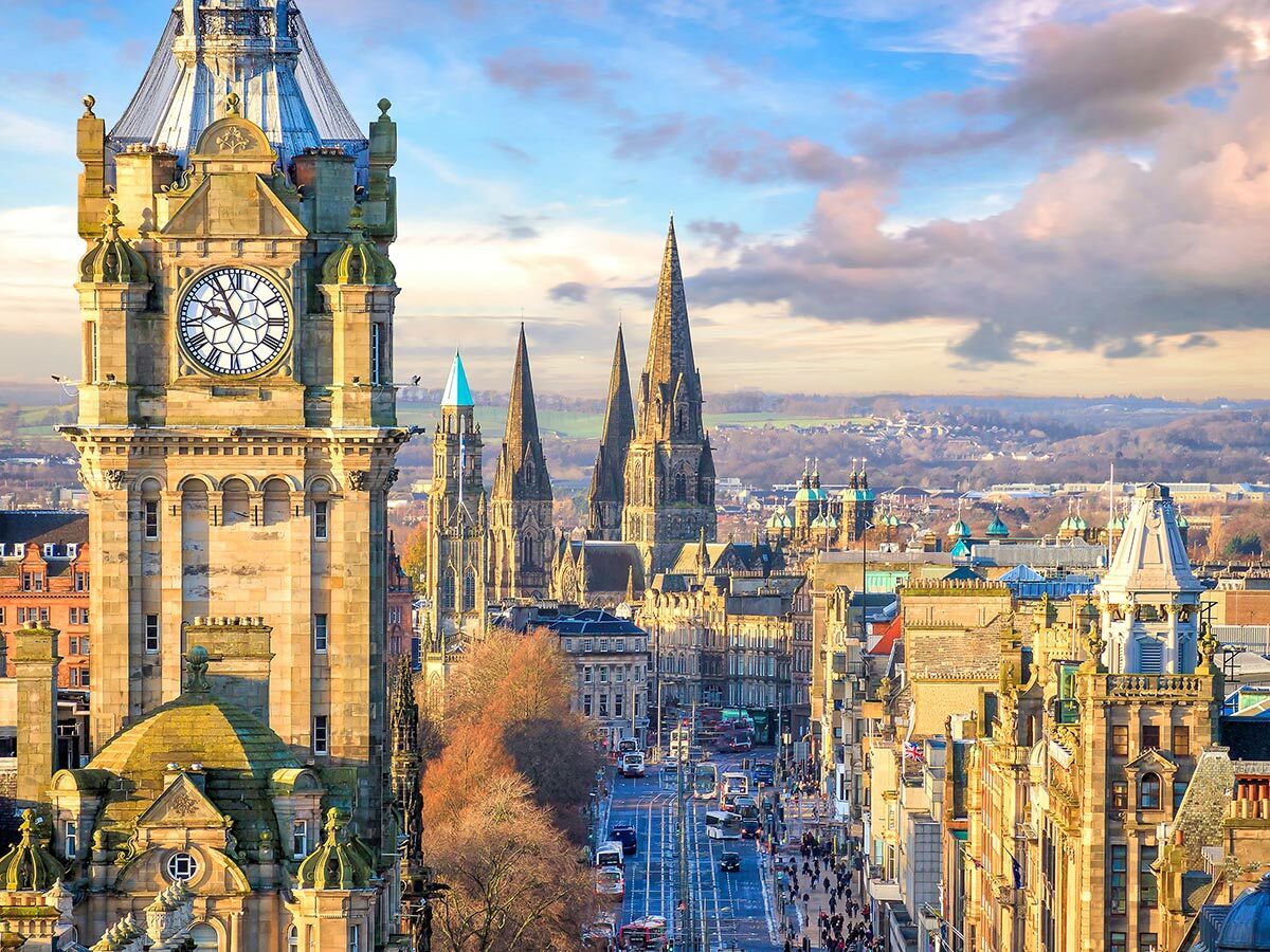 Panoramablick auf Edinburghs belebte Princes Street mit historischem Balmoral Hotel Uhrturm und vielen Kirchtürmen bei blauem Himmel.
