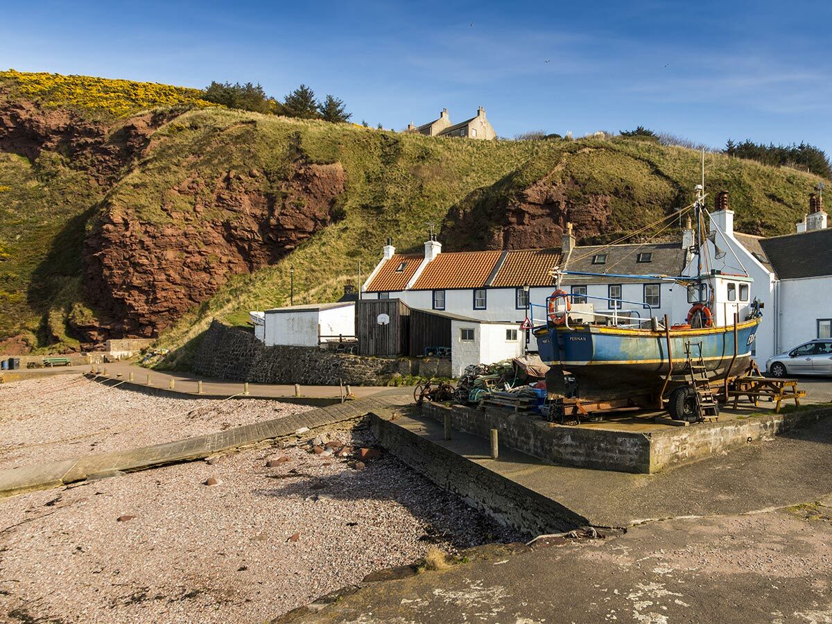 Blaues Fischerboot Pennan auf Trailer an Küste eines schottischen Dorfes, umgeben von Häusern, Strand und Felsenhügel.