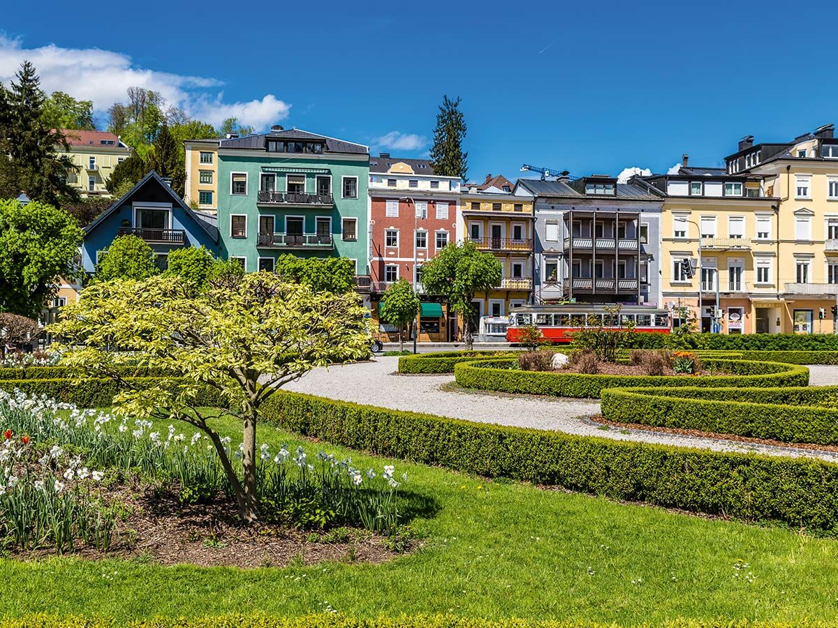 Parkanlage mit Baum, Blumen und Hecken in Gmunden, Österreich. Im Hintergrund bunte Gebäude und eine rote Straßenbahn.