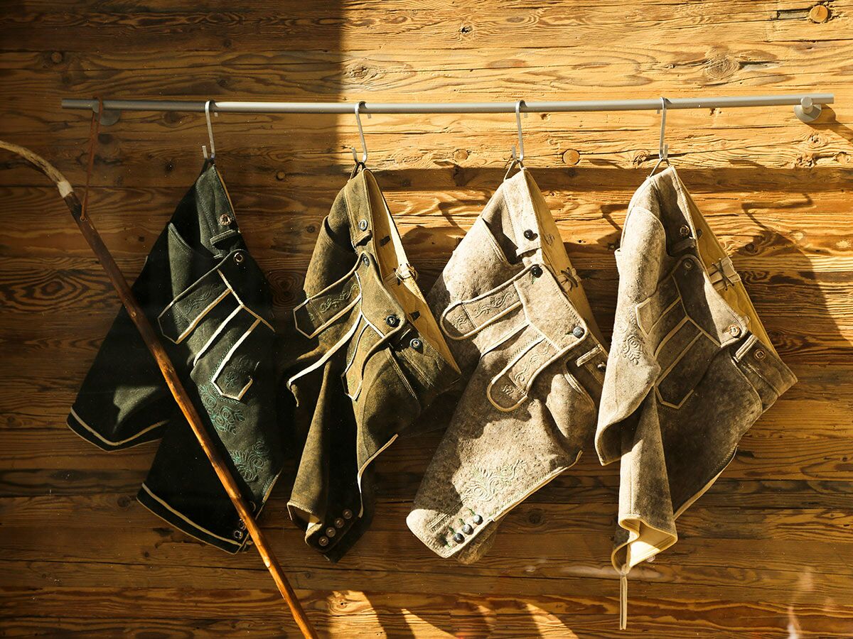 Vier traditionelle, bestickte Lederhosen und ein Holzstock hängen an einer rustikalen Holzwand.
