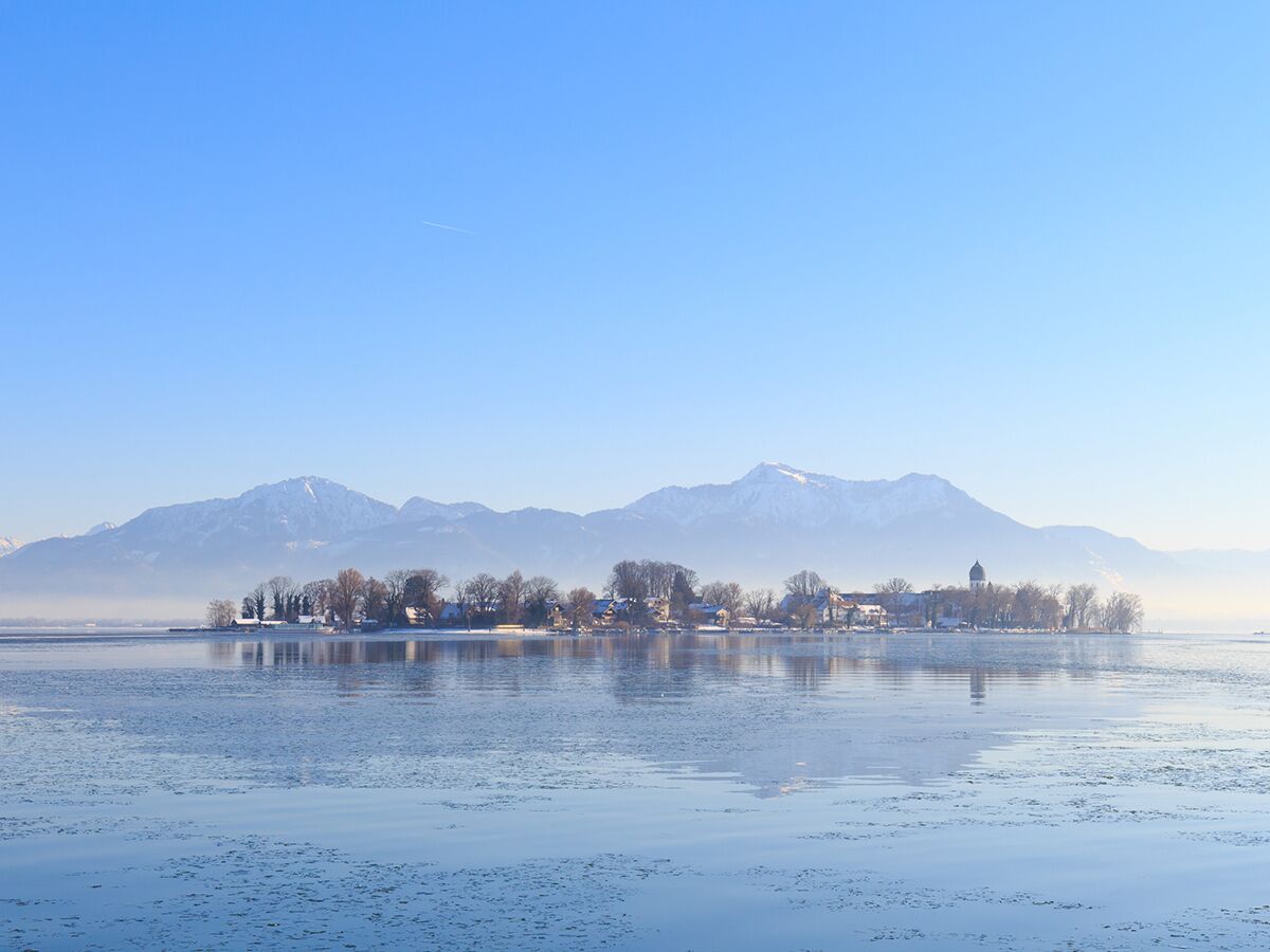 Verschneite Insel mit Dorf und Kirche in einem eisbedeckten See, umgeben von schneebedeckten Bergen unter blauem Himmel.