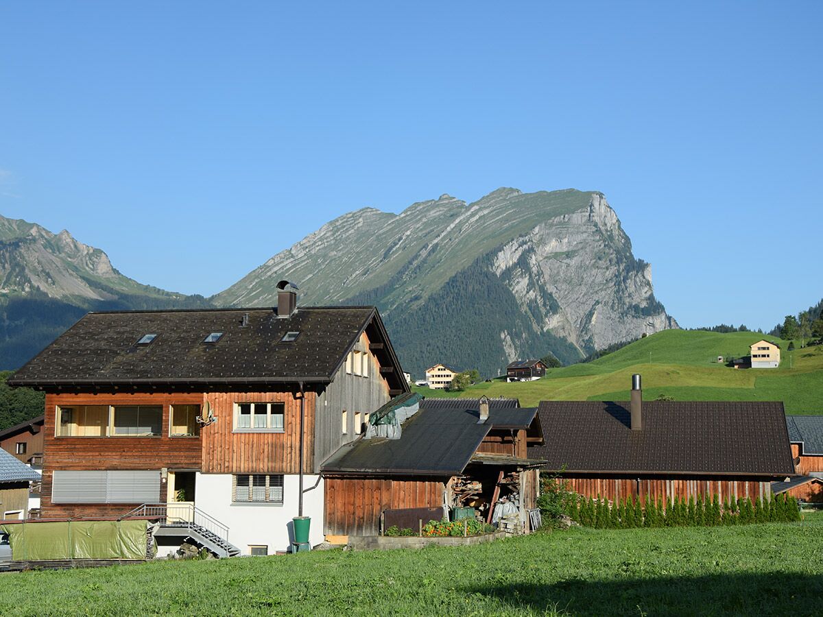 Traditionelle Holzhäuser in einem sonnigen Alpendorf mit grünen Wiesen und majestätischen Bergen im Hintergrund unter blauem Himmel.