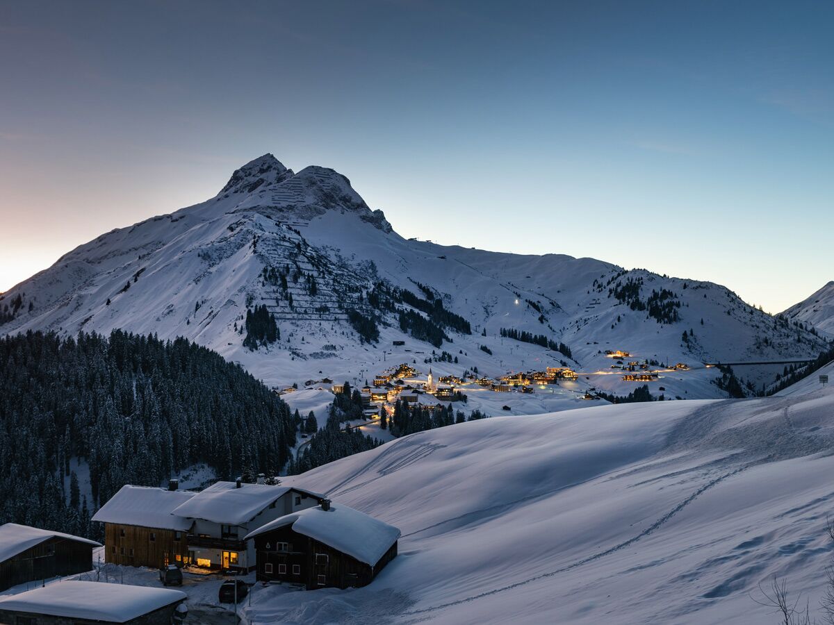 Verschneite Alpenlandschaft bei Dämmerung mit einem beleuchteten Dorf, umgeben von Bergen und warm leuchtenden Häusern.