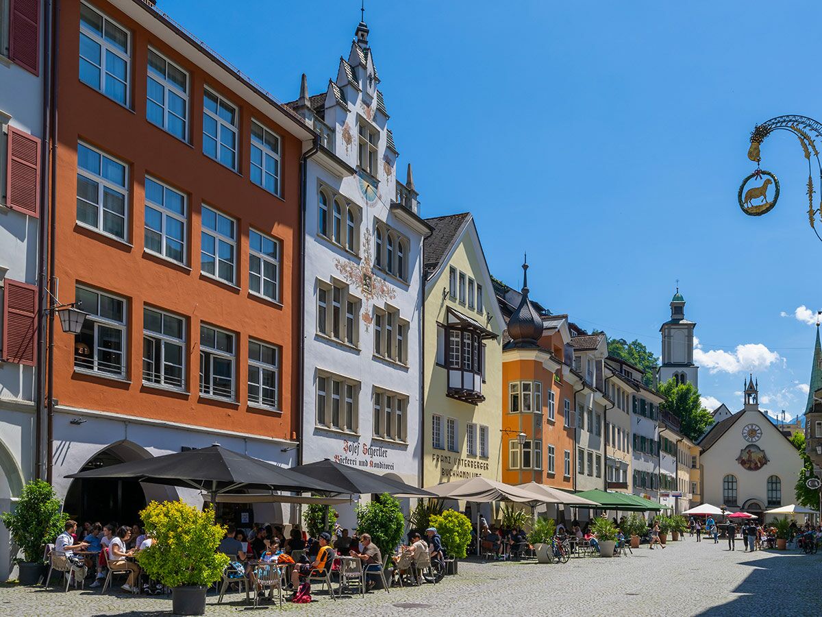 Sonniges Appenzell: Menschen genießen Speisen in belebtem Straßencafé vor historischen Häusern, Kirchturm und goldenem Schafsschild.