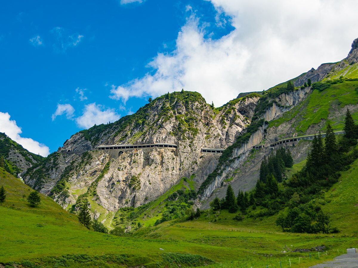 Malerische Berglandschaft mit gewundener Straße, die durch Lawinenschutzgalerien und Tunnel führt, umgeben von grünen Hängen und blauem Himmel.