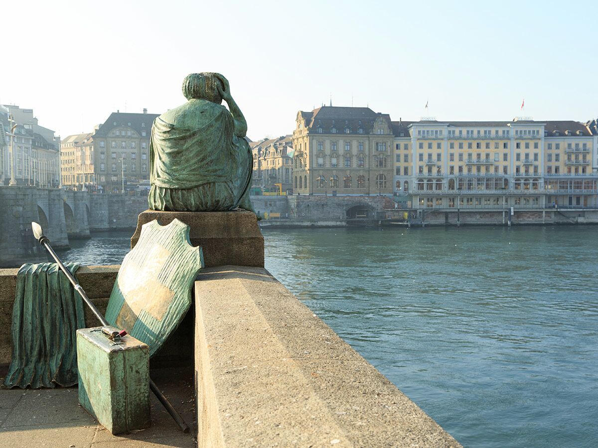 Bronze Statue einer nachdenklichen Figur am Flussufer in Basel, mit Speer und Schild, Blick auf historische Gebäude und Brücke.