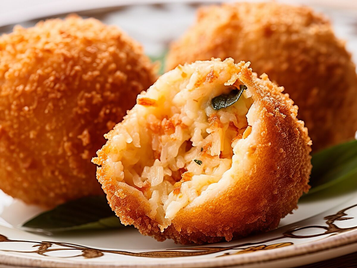 Drei goldbraun frittierte Arancini auf einem Teller, einer aufgeschnitten mit Reis, Tomaten und Kräuterfüllung.