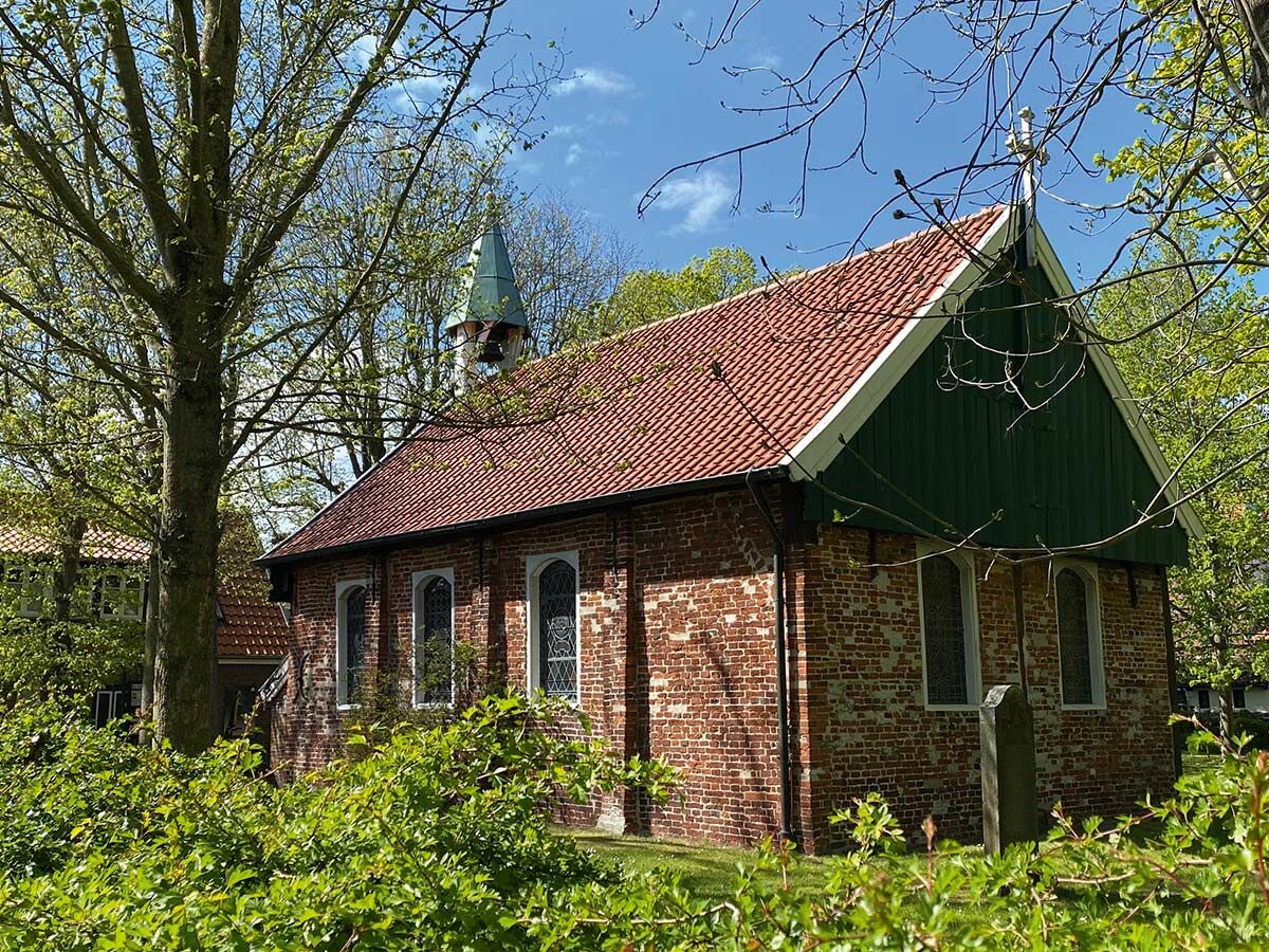 Historische Backsteinkirche mit rotem Ziegeldach, grünem Giebel, Turm und Bäumen unter blauem Himmel. Friedliche Szene.