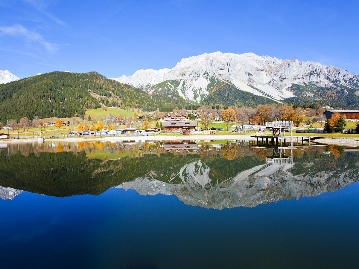 Panorama eines Herbsttages am Bergsee mit schneebedeckten Alpen, bewaldeten Hängen, einem Dorf und klaren Wasserspiegelungen.