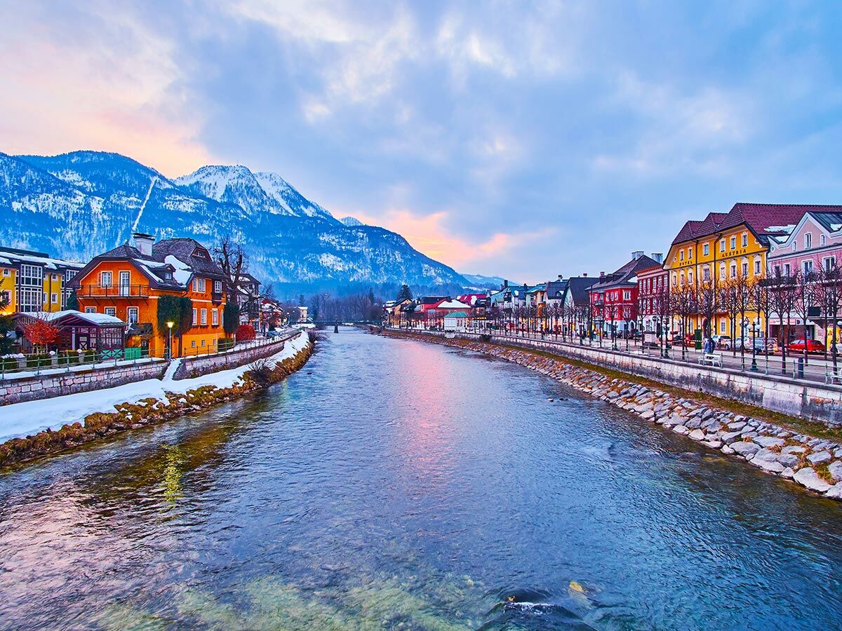 Panorama von Bad Aussee im Winter mit bunten Häusern entlang des Flusses Traun und schneebedeckten Bergen im Hintergrund.