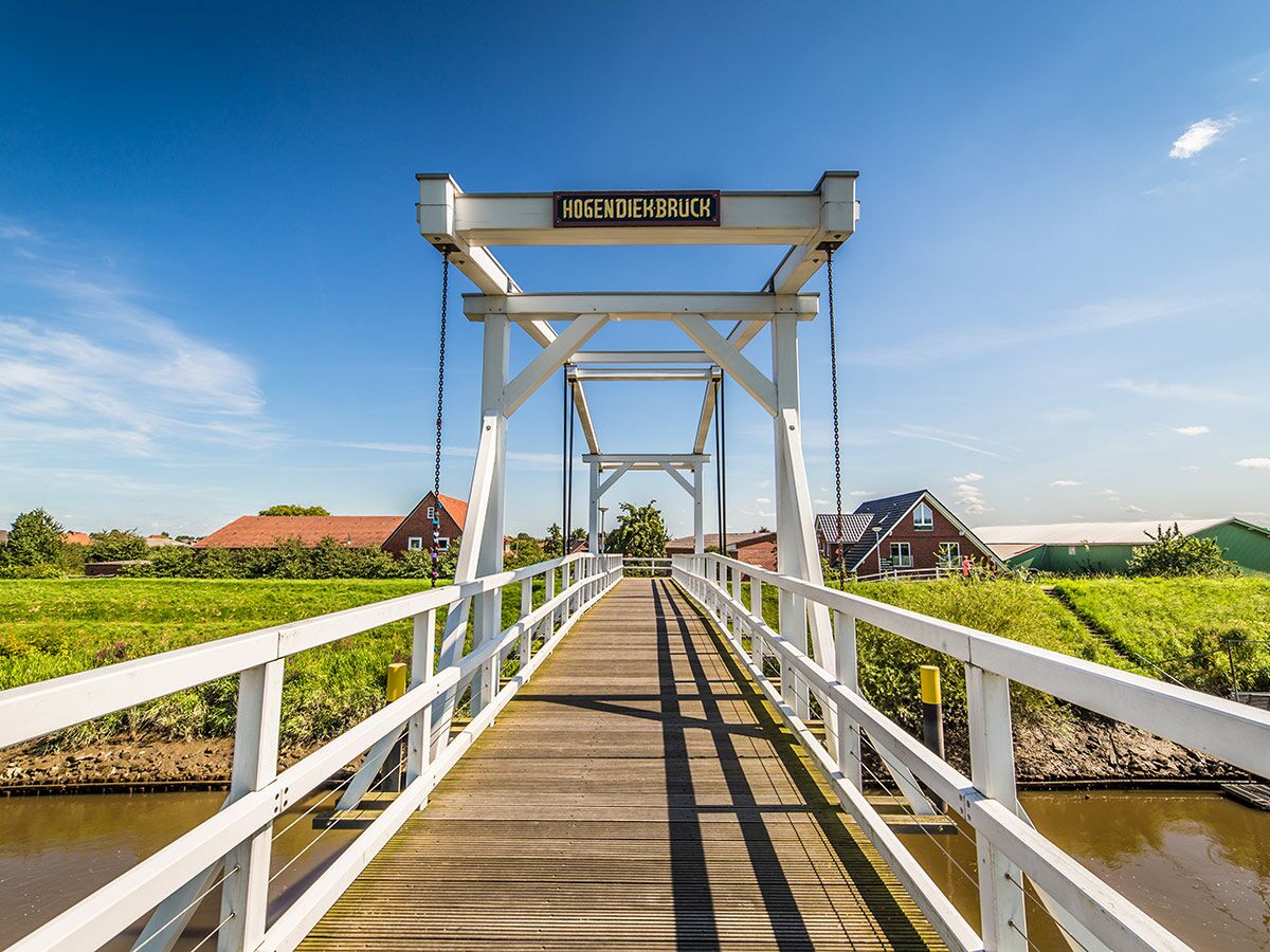 Weiße Fußgängerbrücke HOGENDIEKBRUCH über einen Fluss bei sonnigem Wetter, mit grünen Ufern und Häusern im Hintergrund.