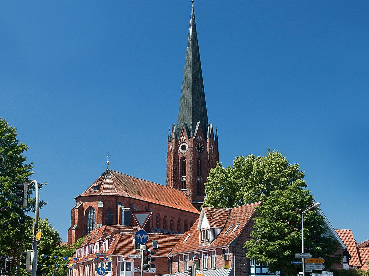 Majestätische Backsteinkirche mit spitzen Turm, Uhr und roten Dächern unter blauem Himmel in einer historischen norddeutschen Stadt.