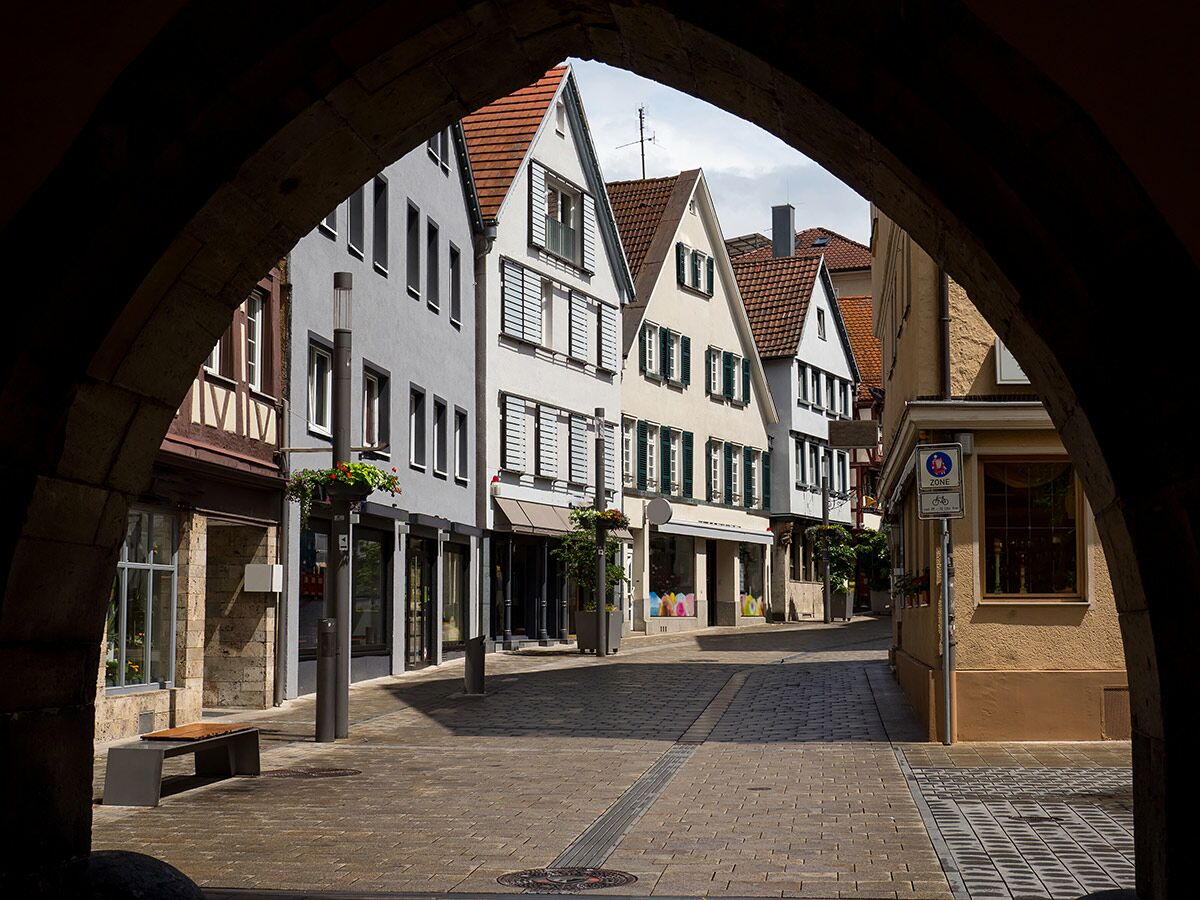 Blick durch dunklen Torbogen auf sonnige, gepflasterte Altstadtgasse mit historischen Häusern, Geschäften und Blumenampel.