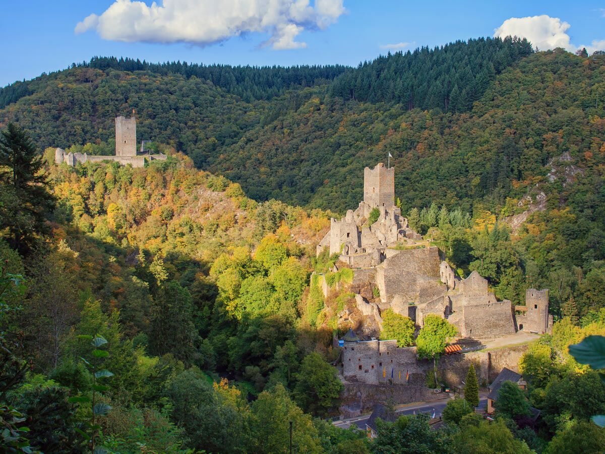Panorama von zwei mittelalterlichen Burgen auf bewaldeten Hügeln im herbstlichen Tal, unter blauem Himmel mit Wolken.