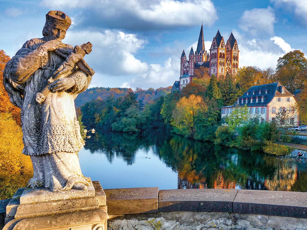 Blick auf den Limburger Dom im Herbst, über die Lahn mit einer Statue im Vordergrund und bunten Bäumen.